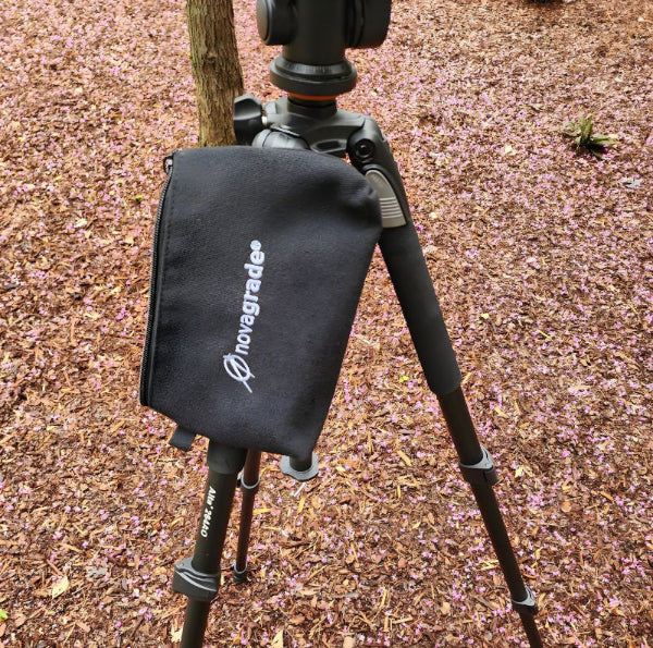 Camera tripod with a black bag featuring 'Novagrade' logo on a ground covered with brown leaves.